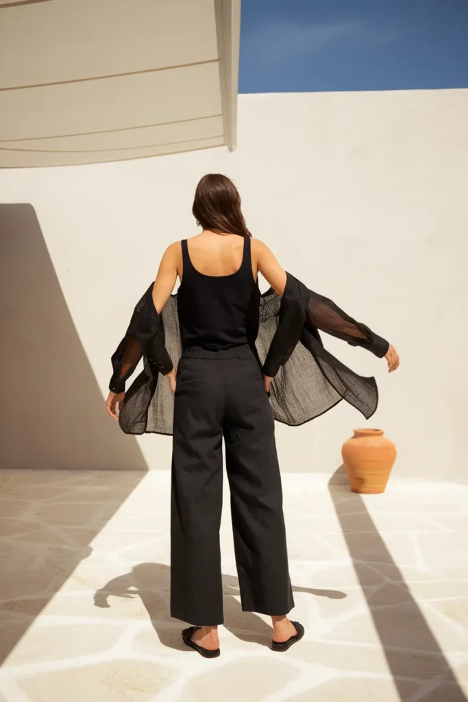 Woman in black linen trousers and a sheer overshirt on a sunlit terrace, summer goth capsule outfit.