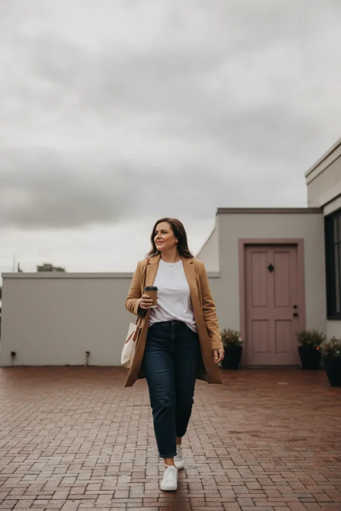 Plus-size woman in a camel longline blazer, white tee, and straight-leg jeans walking on a city sidewalk