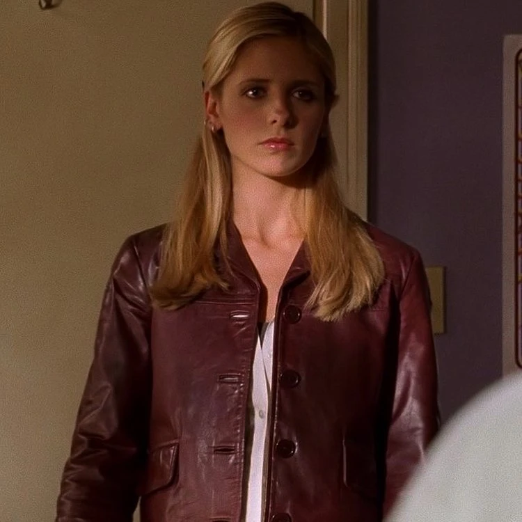 A blonde woman with a serious expression wears a maroon leather jacket over a white top, standing in a hallway between beige and lavender walls.