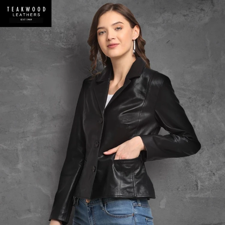 A woman poses against a grey textured backdrop wearing a fitted black leather blazer with notch lapels and button closures, paired with blue jeans, in a Teakwood Leathers product photo.