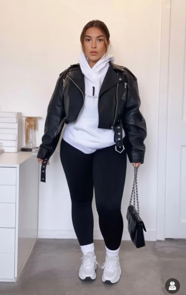 A woman poses indoors wearing a cropped black leather jacket over a white graphic hoodie, black leggings, white sneakers, and a black quilted chain-strap handbag.