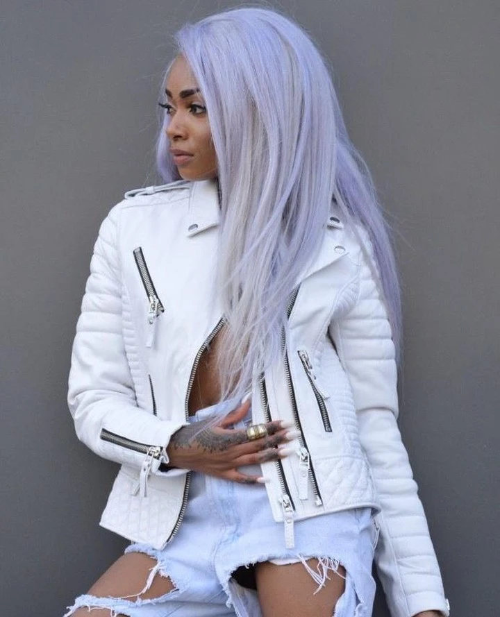 A woman with long lavender hair wears a white leather moto jacket with silver zippers, left partially open, paired with light-wash distressed denim shorts, against a grey backdrop.